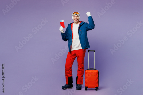Traveler winner man wear blue windbreaker jacket ski goggles mask hat hold passport ticket bag isolated on plain purple background. Tourist travel abroad in free time rest getaway. Air flight concept. #712123873