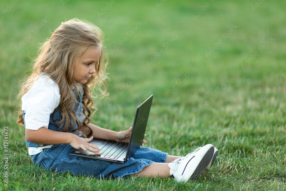 Little girl sitting on the grass with laptop in summer. Happy Caucasian girl child smiling, looking and using at laptop computer in park. Learning online education concept. Little girl sitting on the grass with laptop in summer. Happy Caucasian girl child smiling, looking and using at laptop computer in park. Learning online education concept.