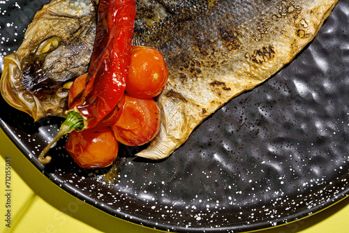 Fried sea bass with baked tomatoes in a black plate on a yellow background
