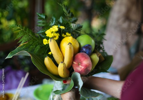 Making of bouquet of fresh ripe fruits, gift for lover.  Hand holding a beautiful fruit bouquet with peach, bananas, apples, flowers. St. Valentine's gift idea. 