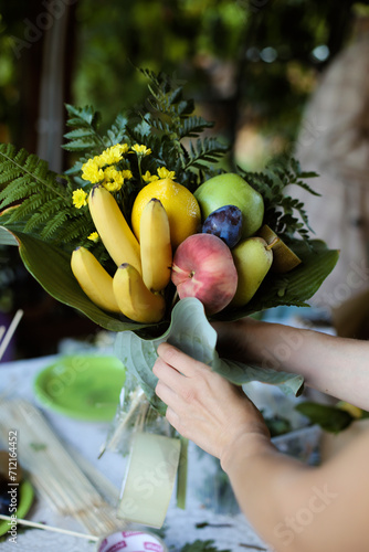 Making of bouquet of fresh ripe fruits, gift for lover.  Hand holding a beautiful fruit bouquet with peach, bananas, apples, flowers. St. Valentine's gift idea. 