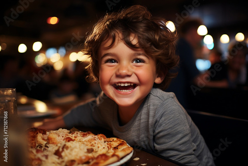 A happy laughing curly-haired boy at the table is eating fresh pizza  holding food in his hands. A joyful atmosphere in the pizzeria