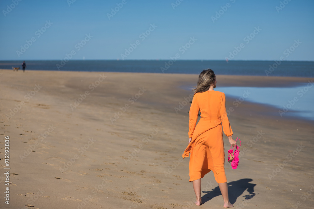 Young and beautiful woman in an orange dress and pink heels in her hand, walking barefoot on the beach in solitude. Concept beauty, fashion, trend, loneliness.