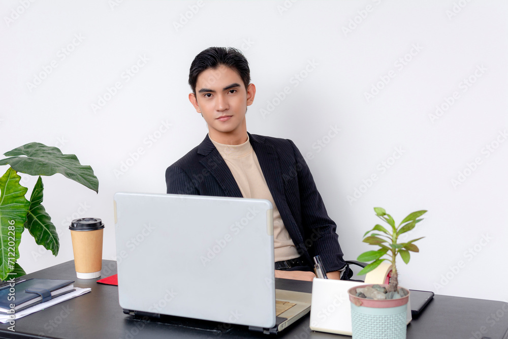 A confident young Asian businessman in a suit working on a laptop at his tidy office desk with coffee and plants. A confident young Asian businessman in a suit working on a laptop at his tidy office desk with coffee and plants.