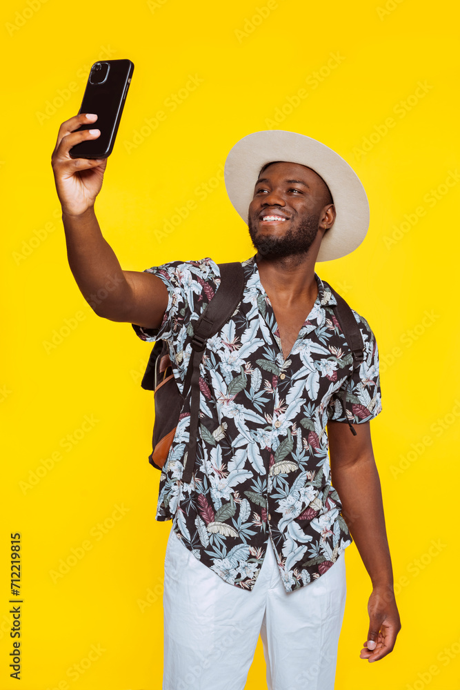 Young happy smiling bearded male student of african american ethnicity wears floral hawaiian shirt, backpack and stylish hat, doing selfie shot on mobile smart phone isolated on yellow background. Young happy smiling bearded male student of african american ethnicity wears floral hawaiian shirt, backpack and stylish hat, doing selfie shot on mobile smart phone isolated on yellow background.