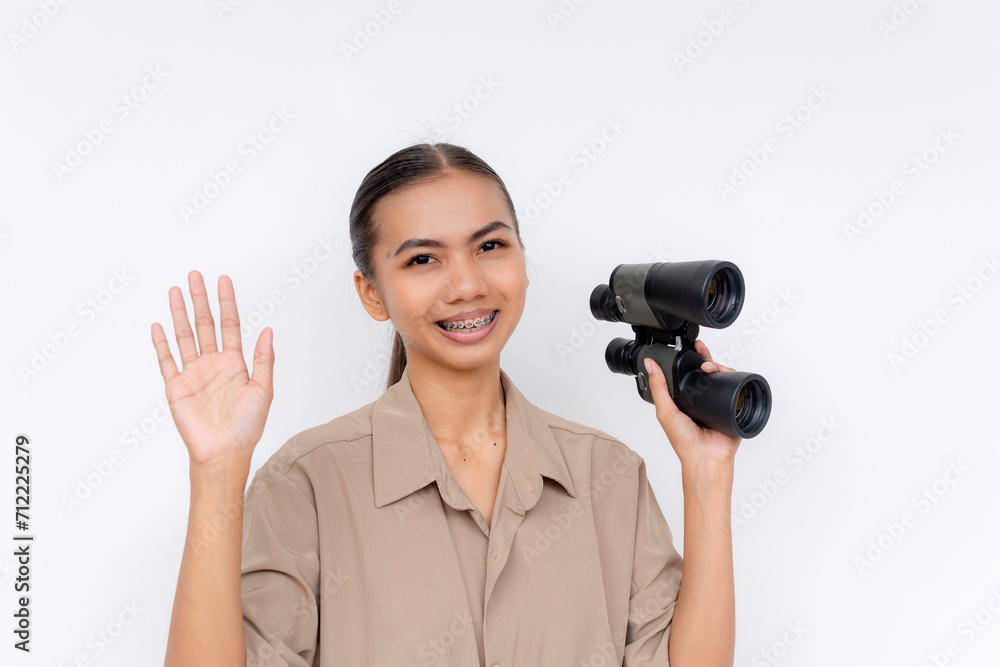 Cheerful young Filipino woman with braces waving hello while holding binoculars against a white background. Cheerful young Filipino woman with braces waving hello while holding binoculars against a white background.