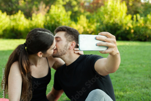 Pareja heterosexual caucasica enamorados haciendose un selfie en el cesped del parque posando besándose photo