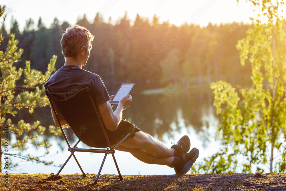 A man is reading an e-book on the shore of a forest lake at sunset in the evening. - obrazy, fototapety, plakaty 