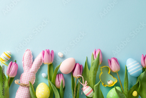 Family Easter setup. Top view of lively eggs, playful cookie cutter, charming bunny, and tulips against a pastel blue background. Ample space for text or promotions #712238631