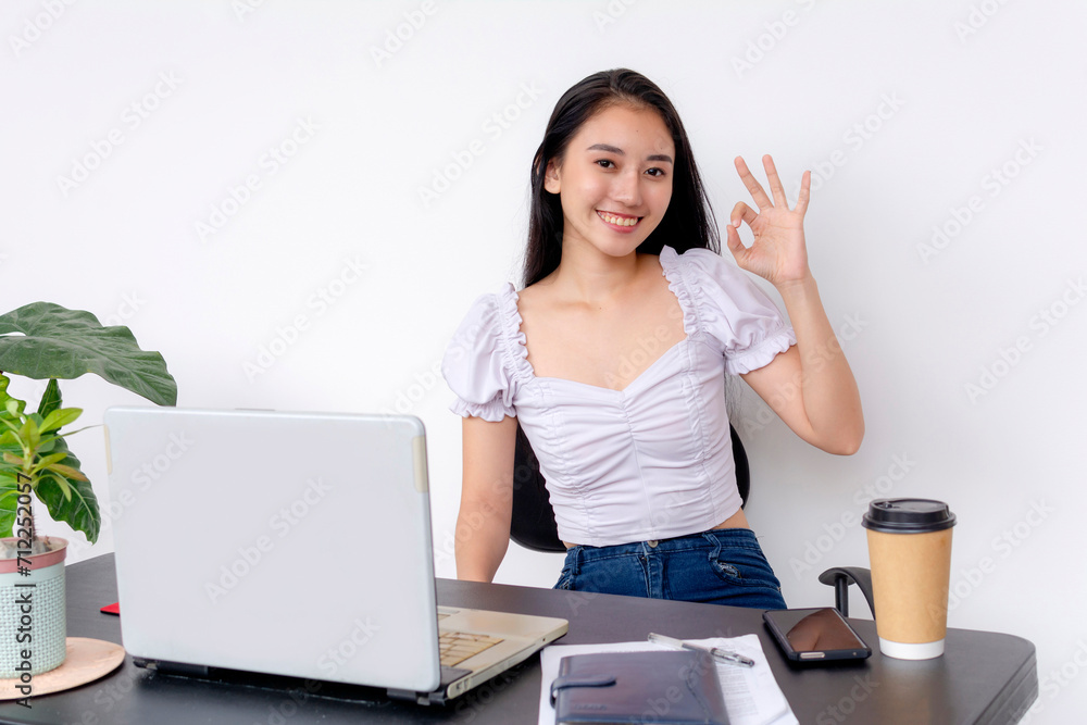 A studious young female student smiling and giving an ok sign while studying at her desk with a laptop and coffee. A studious young female student smiling and giving an ok sign while studying at her desk with a laptop and coffee.