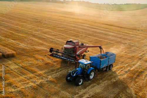 Combine reaper pours wheat grains into tractor trailer in empty field aerial view. Agricultural machinery transports gathered crop at sunset photo