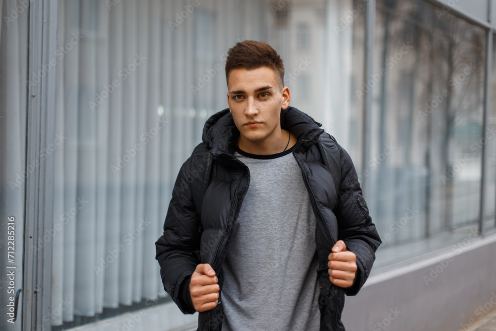 handsome fashionable young man with hairstyle in a winter down jacket and T-shirt stands near a building on the street handsome fashionable young man with hairstyle in a winter down jacket and T-shirt stands near a building on the street