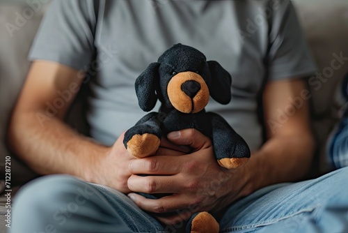 Middle aged man holds toy black brown stuffed dog in hand wearing gray T shirt indoors focused photo