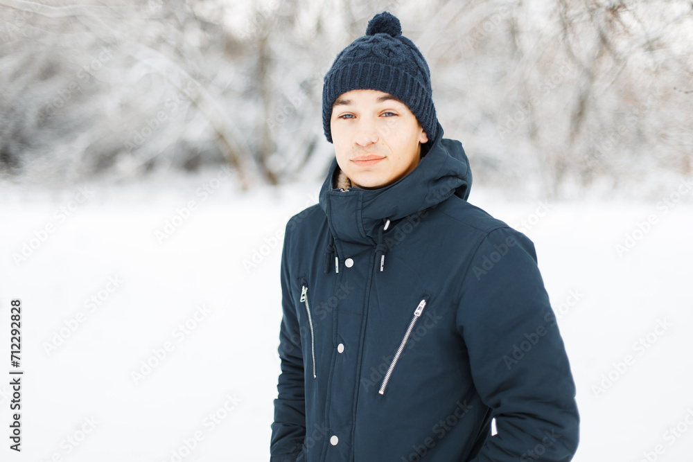 Handsome young guy with a knitted hat in a fashion winter jacket against the background of a snowy park outdoors. Handsome young guy with a knitted hat in a fashion winter jacket against the background of a snowy park outdoors.