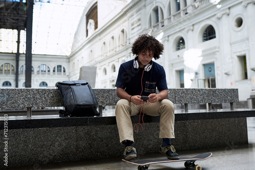 Hipster boy listening to music sitting on bench with feet on skateboard.