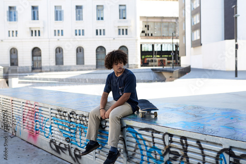 Boy with curly hair sitting beside his skateboard.