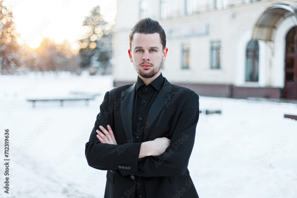 Stylish fashionable man model in a elegant black suit with a blazer and a shirt stands in the city on the snowy winter day at sunset Stylish fashionable man model in a elegant black suit with a blazer and a shirt stands in the city on the snowy winter day at sunset