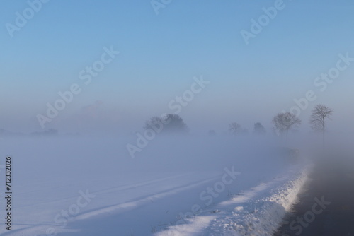 Trees on the horizon are barely visible during sunrise on a cold winter day with mist and snow.