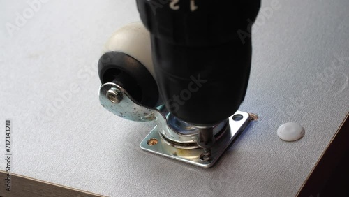 The screwdriver tightens the screw. An electric handheld screwdriver drives a black hardened screw into a wooden slab and secures a furniture wheel. Furniture assembly workshop. Close-up of screw