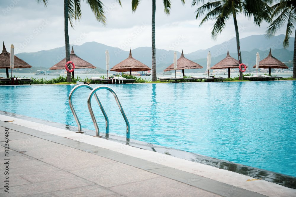 Luxury ocean view tropical resort swimming pool with stainless steel handrail, row of beach umbrella thatched with palm leaves, beach and mountain range background in Nha Trang, Vietnam