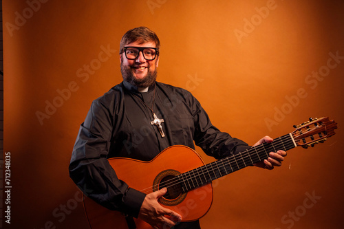 Portrait of a priest with a crucifix and black shirt playing the guitar.
