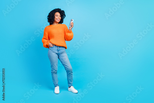 Full body photo of pretty young girl hold device chatting comment dressed stylish knitted orange outfit isolated on blue color background