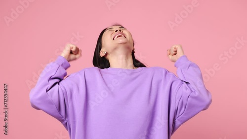 Excited jubilant overjoyed happy young woman of Asian ethnicity wear purple sweatshirt do winner gesture celebrate clench fists say yes isolated on plain pastel light pink background studio portrait