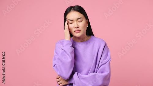 Sick ill sad tired young woman of Asian ethnicity wear purple sweatshirt put hands on head rub temples having headache suffering from migraine feel bad seedy isolated on plain pastel pink background photo