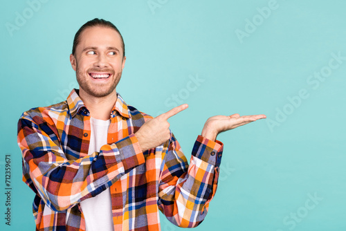 Portrait of toothy beaming guy wear flannel shirt indicating look at product on arm empty space isolated on turquoise color background