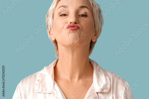Beautiful mature woman in pajamas doing face building exercise on blue background  closeup