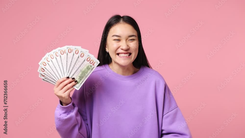 Happy young woman of Asian ethnicity wearing purple sweatshirt hold in hand fan of cash money in dollar banknotes doing winner gesture celebrate isolated on plain pastel light pink background studio Happy young woman of Asian ethnicity wearing purple sweatshirt hold in hand fan of cash money in dollar banknotes doing winner gesture celebrate isolated on plain pastel light pink background studio