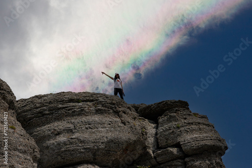 A young girl at the peak of a mountain and a rainbow in the sky.