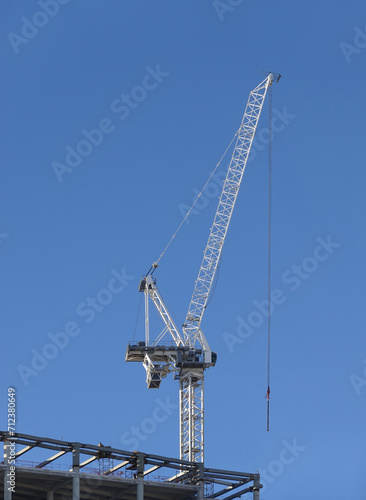 Hoisting tower crane on the top section being constructed of modern high skyscraper building against blue cloudless sky