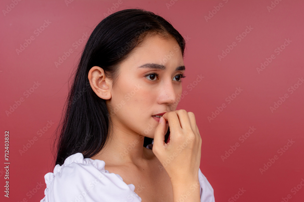 An anxious and jittery young Asian woman biting her nail in an attempt to calm down. Portrait shot against a vibrant red background. An anxious and jittery young Asian woman biting her nail in an attempt to calm down. Portrait shot against a vibrant red background.