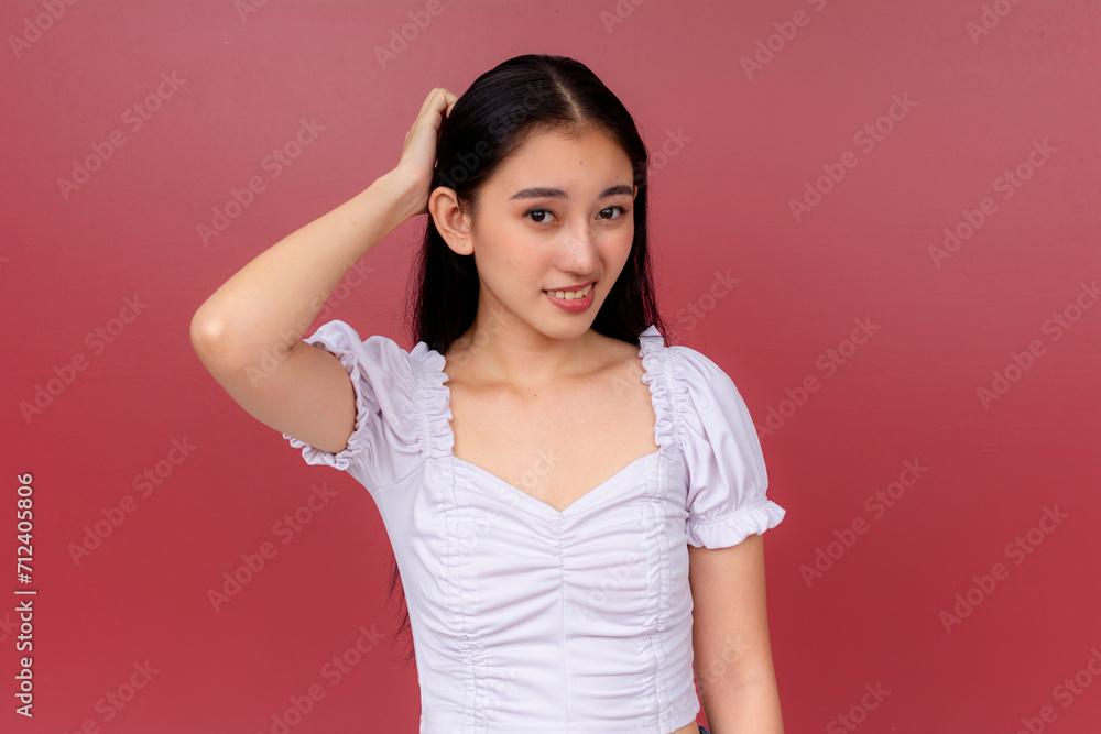 Fototapeta premium A radiant Asian woman in a white blouse smiles charmingly while scratching her head in ignorance after misjudging someone. Standing before a vivid red backdrop.