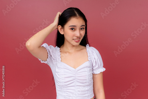 A radiant Asian woman in a white blouse smiles charmingly while scratching her head in ignorance after misjudging someone. Standing before a vivid red backdrop. photo