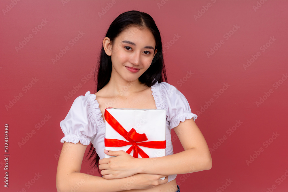 Cheerful Asian woman in white blouse holding a present with red ribbon, conveying happiness and celebration on a red backdrop. Cheerful Asian woman in white blouse holding a present with red ribbon, conveying happiness and celebration on a red backdrop.