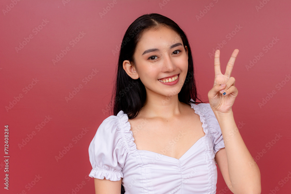 Cheerful Asian woman smiling and making a peace sign with her hand against a vibrant red background. Cheerful Asian woman smiling and making a peace sign with her hand against a vibrant red background.