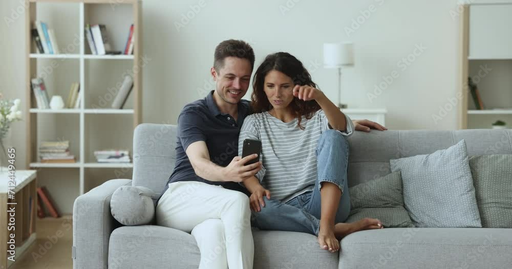 Positive married couple using smartphone together at home, sitting close on sofa, discussing online application, taking selfie, talking on video call, relaxing on couch with digital gadget Positive married couple using smartphone together at home, sitting close on sofa, discussing online application, taking selfie, talking on video call, relaxing on couch with digital gadget