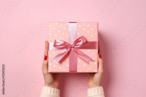 Hands of young woman holding beautiful gift box with pink ribbon on pink background flat lay. Surprise gift in hands close-up. Congratulations on the holiday, March 8, Mother's Day