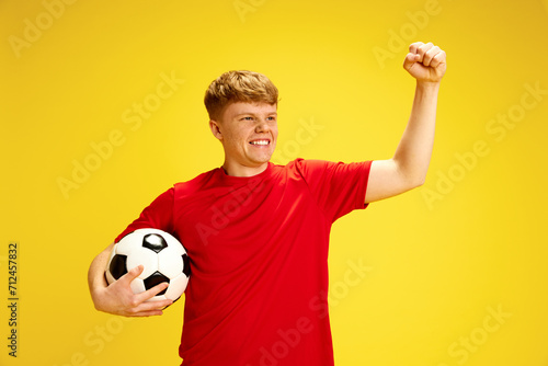 Promotional material for community soccer league. Young man in red shirt holding soccer ball and making victory gesture against yellow background. Active lifestyle, youth, hobby and emotions concept photo