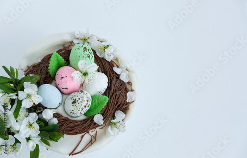 Easter cake is decorated like nest with colorful eggs, branch of spring white flowers on white background with copy space. Easter postcard, spring background.