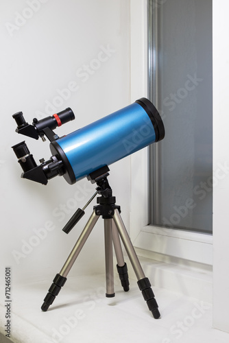 Blue catadioptric astronomical telescope on a tripod in the room. The telescope is on the windowsill and pointed out the window