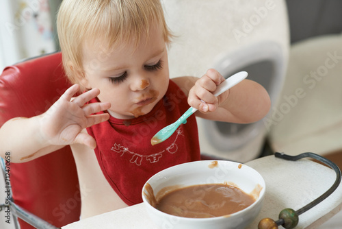 High chair  eating and baby with spoon in a house for nutrition  diet and fun while playing. Food  messy eater and boy kid at home with meal for child development  fiber or vitamins while learning