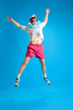 Full-length image of young happy, positive man in summer trip, jumping, having fun against blue studio background. Concept of emotions, youth, leisure time, summer vacation, travelling