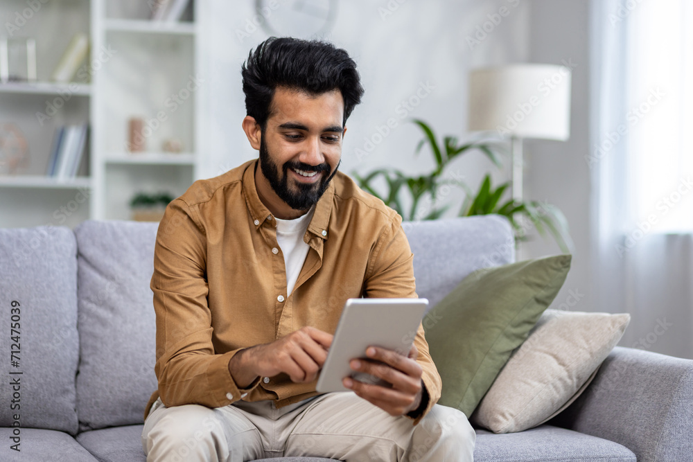 Indian man smiles while browsing on a tablet, comfortably seated on a sofa in a well-lit modern apartment living room.