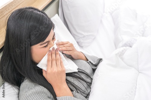 Young woman sick flu caught sneezing in home. holding hand tissue paper. female work in bed rhinitis runnning nose. health problem allergy. girl beautiful asian lying tablet on lap feeling got fever. photo