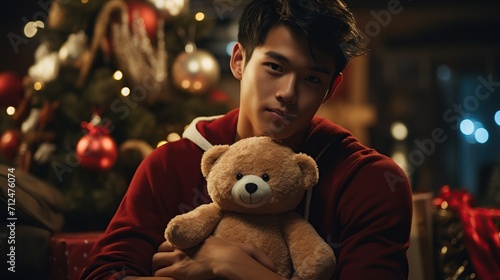 handsome and cute young guy sitting near the Christmas tree and hugging a teddy bear in a santa claus costume