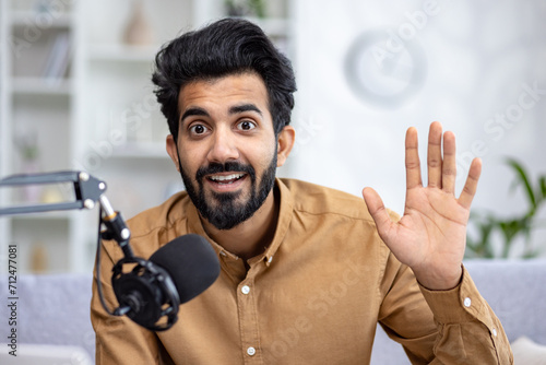 A cheerful young Indian man waving while talking into a microphone, sitting on a sofa in the cozy living room of an apartment. photo