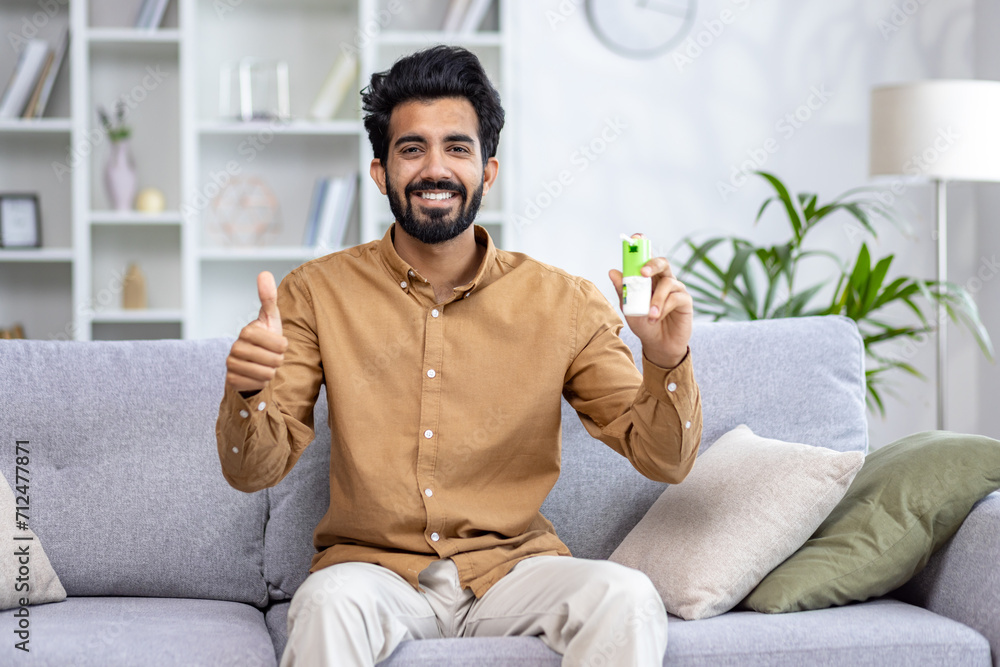 Smiling Indian man sitting comfortably on a couch at home, gesturing thumbs up, showing positivity and approval. Smiling Indian man sitting comfortably on a couch at home, gesturing thumbs up, showing positivity and approval.
